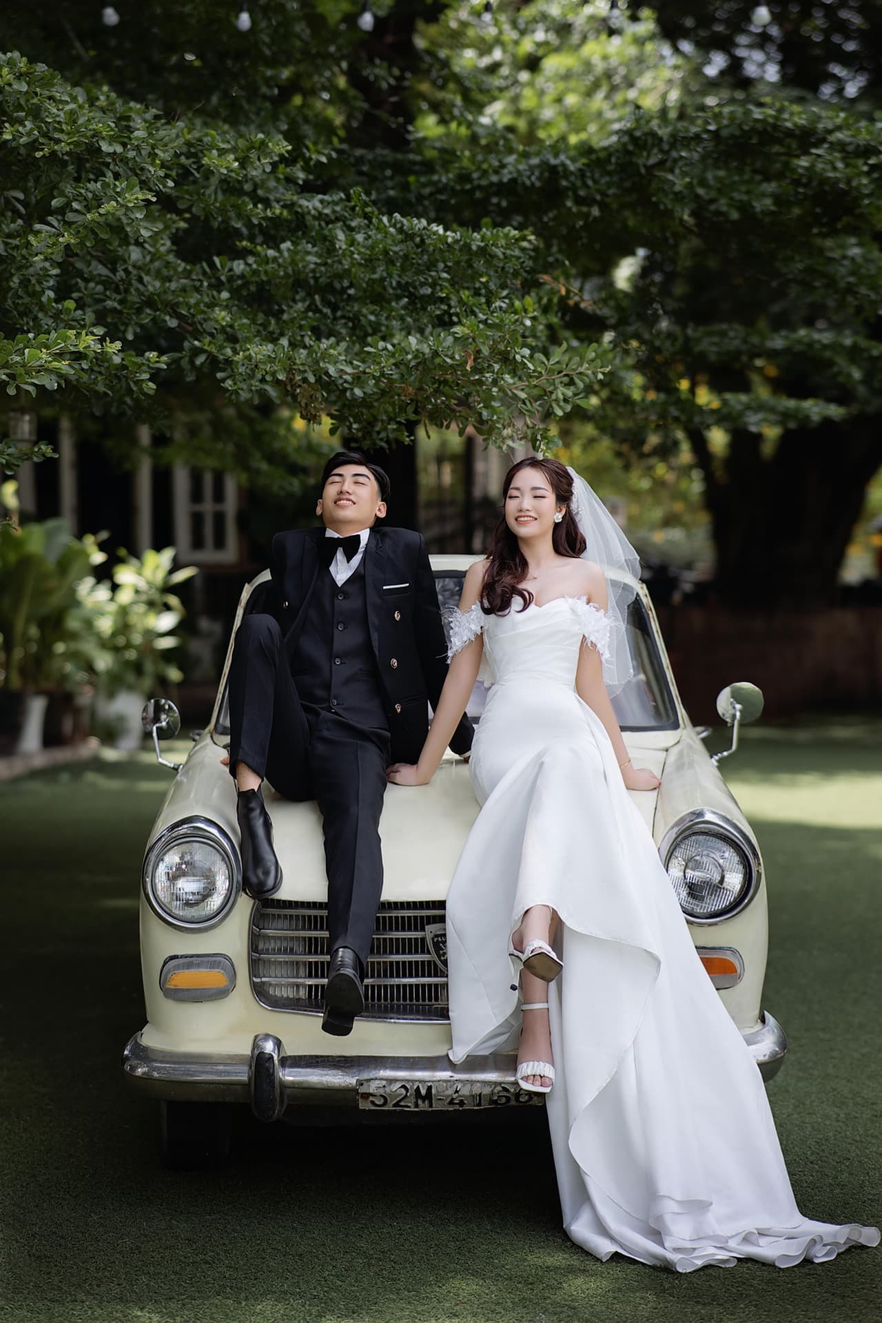 A couple seated on the bonnet of a vintage car under green trees